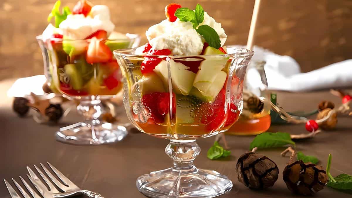 Two glass dessert cups filled with colorful fruit salad, topped with whipped cream and mint leaves. The table is adorned with autumn decor, including dried pinecones and leaves, with a fork placed in the foreground.