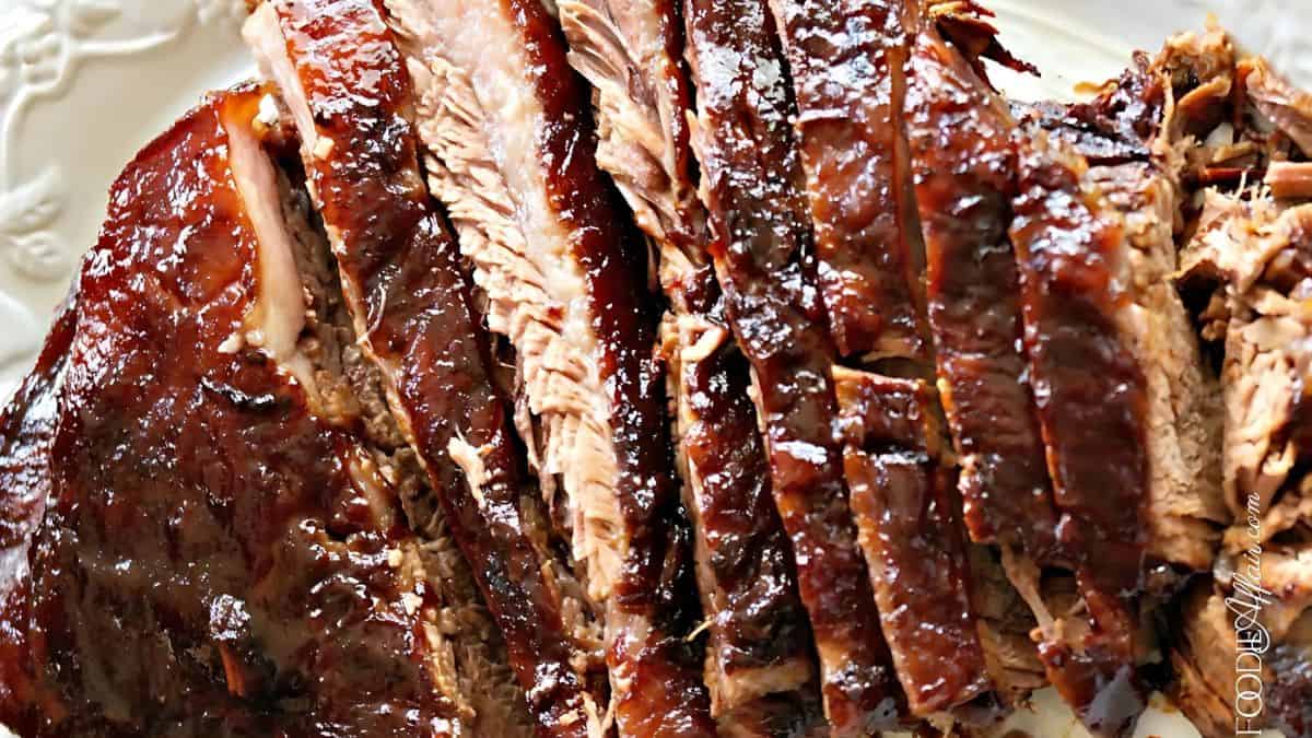 Close-up of sliced, juicy barbecue brisket with a rich, dark glaze on a white plate. The meat is tender and appears well-cooked, showcasing a glossy, caramelized exterior.