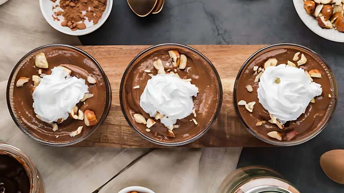 Three bowls of chocolate pudding topped with whipped cream and chopped nuts sit on a wooden board. Surrounding them are ingredients like cocoa powder and almonds, along with a spoon on a dark surface.