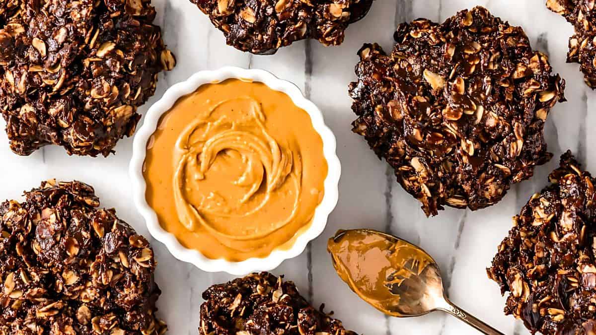 A bowl of creamy peanut butter surrounded by chocolate oat cookies on a marble surface. A spoon with peanut butter is placed next to the bowl. The cookies are studded with oats and dark chocolate chunks.