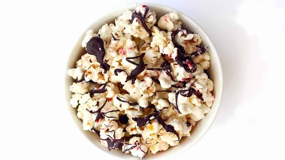 A bowl of popcorn drizzled with dark chocolate and sprinkled with crushed peppermint on a white background.