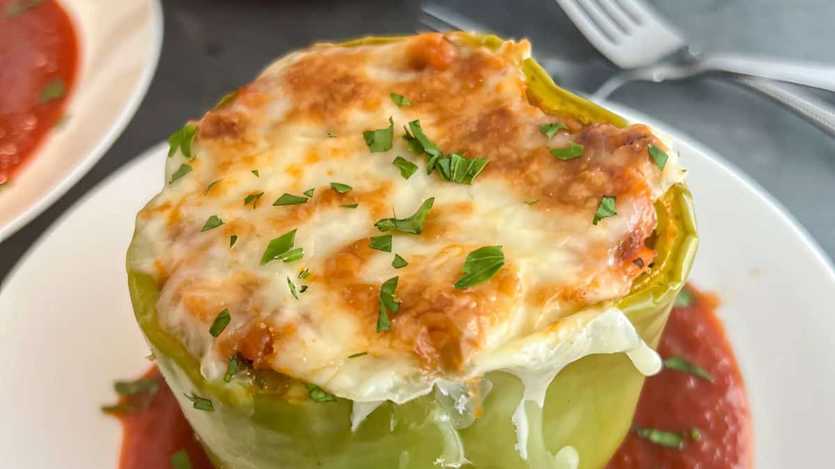 A baked green bell pepper stuffed with melted cheese and garnished with chopped herbs sits on a white plate surrounded by a red sauce. A fork is visible in the background.