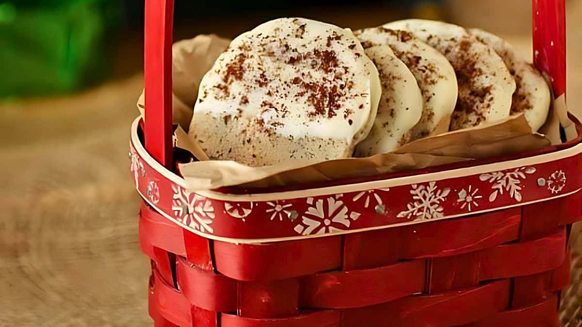 A red woven basket with a snowflake design holds several round, white cookies sprinkled with brown crumbs. The basket is lined with brown paper and is placed on a wooden surface.