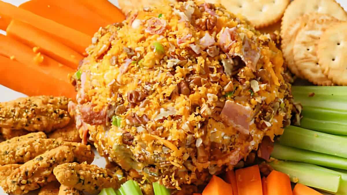 A cheese ball coated with chopped nuts and herbs, surrounded by carrot sticks, celery sticks, round crackers, and seasoned breadsticks on a platter.