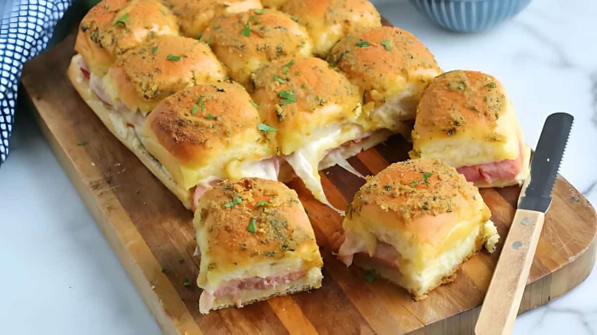 A batch of cheesy ham and cheese sliders on a wooden cutting board. The sandwiches are topped with herbs and a golden crust. One slider is partially cut and separated from the rest with a knife beside it. A napkin and bowl are in the background.