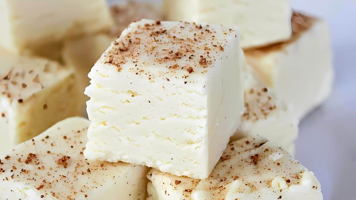 Close-up of creamy white chocolate fudge pieces topped with a light dusting of cocoa powder. The fudge has a smooth texture with some subtle layering, suggesting a rich and creamy consistency. Pieces are stacked attractively on a white surface.