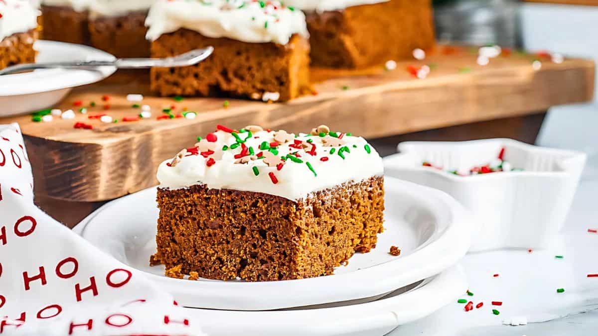 A piece of gingerbread cake with white frosting and festive red, green, and white sprinkles on a white plate. In the background, a wooden board holds more pieces of decorated cake. A napkin with "Ho Ho Ho" is partially visible.