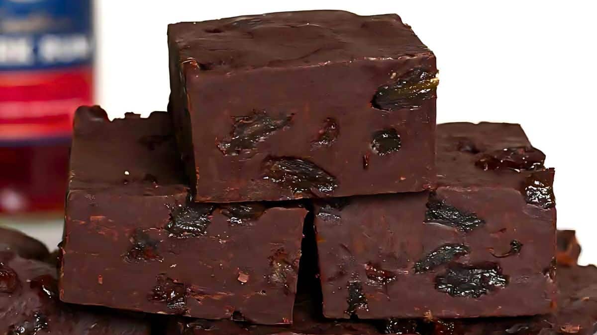 Close-up of a stack of dark chocolate fudge pieces with visible chunks of dried fruit, possibly raisins, embedded in them. A bottle with a partially visible label is in the background.