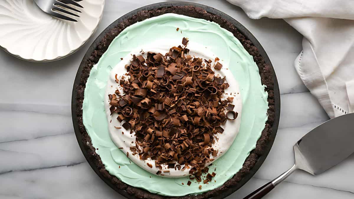 A creamy green pie with a chocolate crust, topped with whipped cream and chocolate shavings. A pie server is on the right, and a dessert plate with a fork is in the top left corner on a marble surface.
