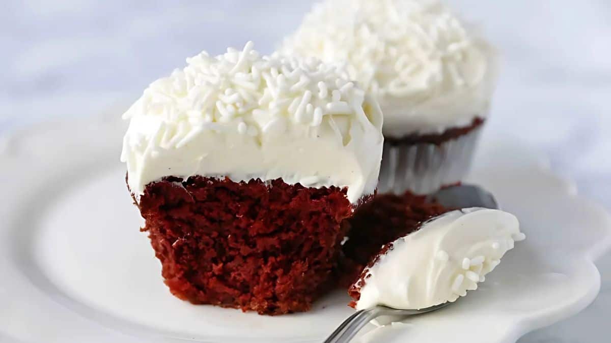 A close-up of a red velvet cupcake with white frosting and sprinkles on a white plate. The cupcake is cut in half, showing its rich red interior, with a spoon next to it holding a piece of the cupcake.