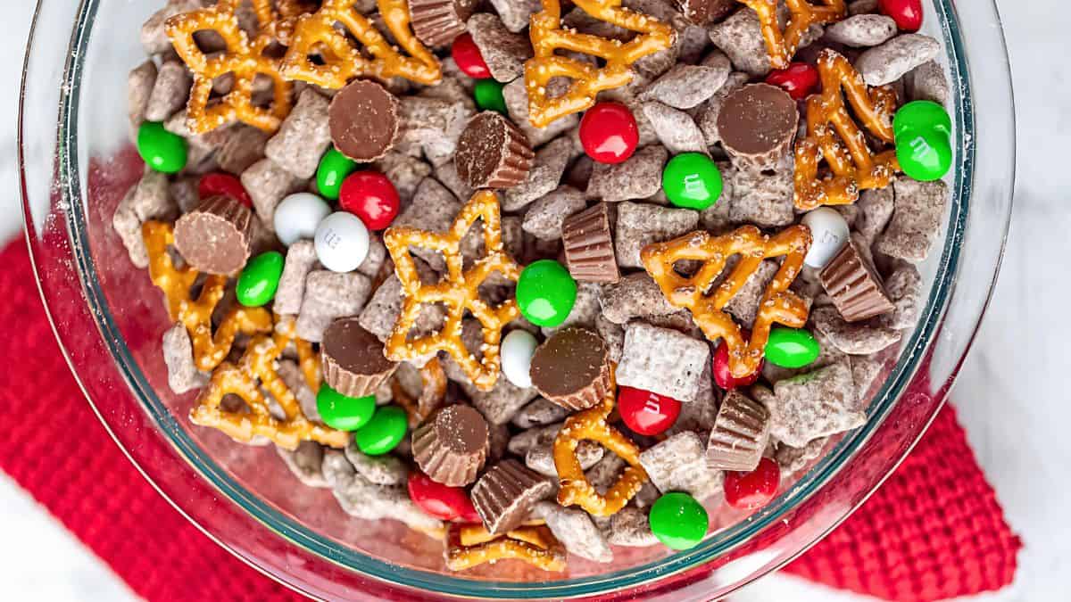 A glass bowl filled with holiday snack mix, including chocolate-covered cereal pieces, red and green candy-coated chocolates, mini peanut butter cups, and star-shaped pretzels, sits on a red textured placemat.