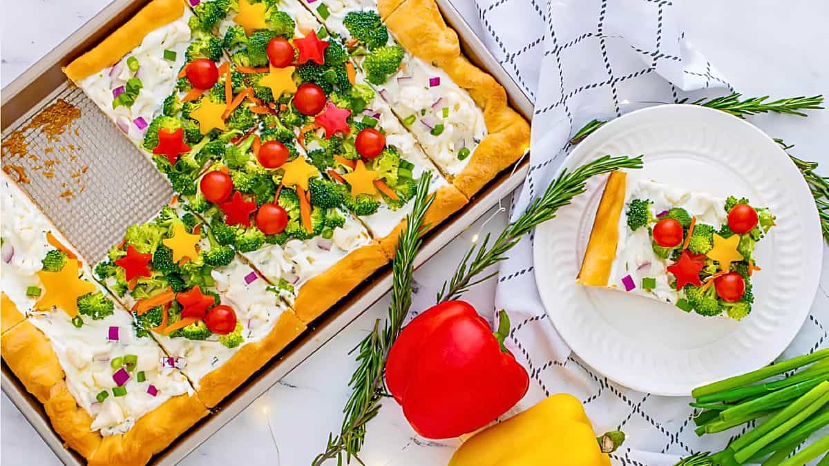 A festive vegetable pizza with cream cheese topping, decorated with broccoli, cherry tomatoes, red onion, and star-shaped bell pepper pieces. One slice is on a white plate. Nearby are sprigs of rosemary and whole bell peppers.