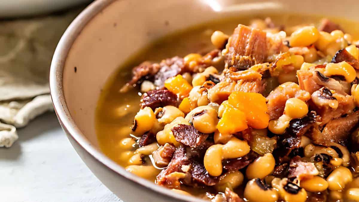 A bowl of savory stew with black-eyed peas, chunks of ham, and diced vegetables in a rich, hearty broth. The dish is served in a rustic, shallow bowl on a light-colored surface.