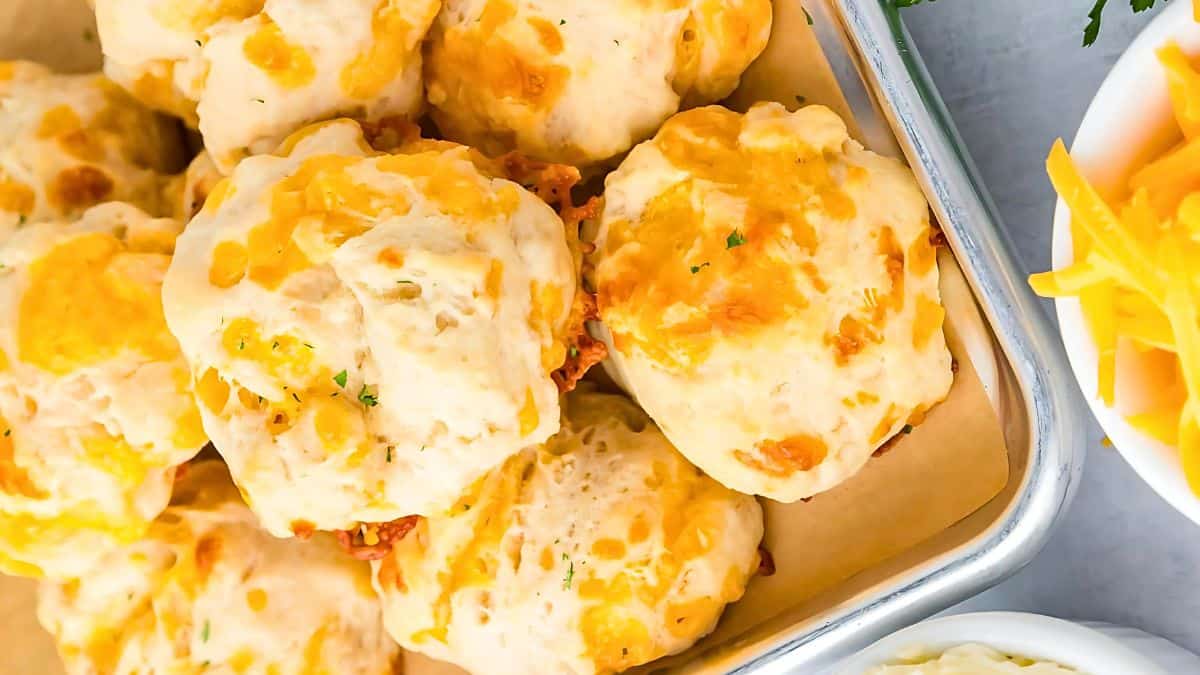 A tray of golden-brown cheddar biscuits, topped with melted cheese and garnished with herbs. Some shredded cheese is visible in a bowl nearby.