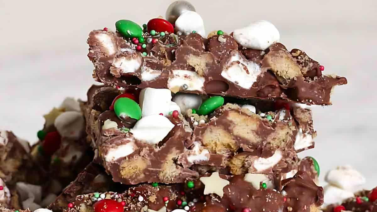 A close-up of stacked rocky road pieces, featuring chocolate, marshmallows, cookie chunks, and colorful candies. The candies are red, green, and silver, with small white star sprinkles adding a festive touch.