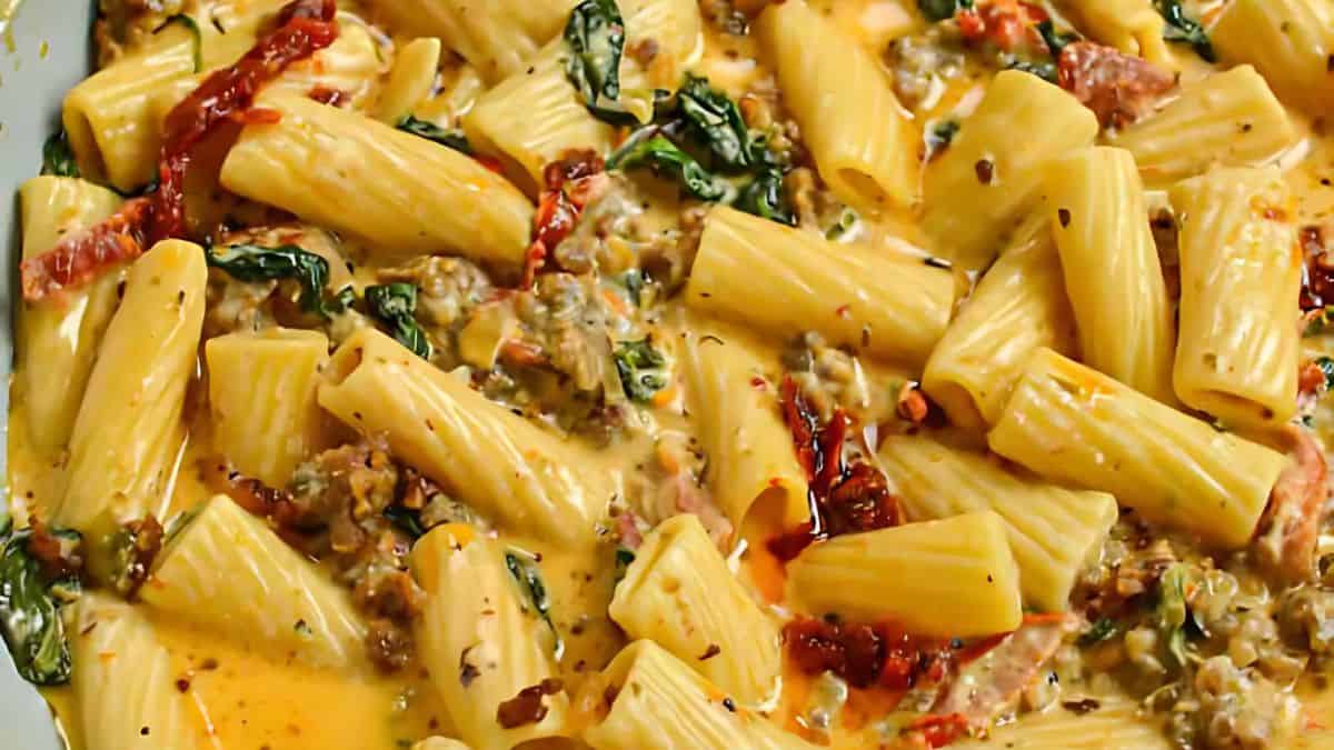Close-up of a creamy pasta dish featuring rigatoni noodles mixed with spinach, sun-dried tomatoes, and a savory sauce, garnished with herbs and spices.