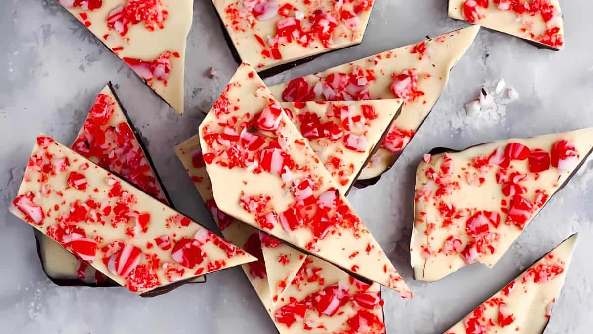 Pieces of white chocolate peppermint bark with crushed candy canes sprinkled on top are spread out on a gray surface.