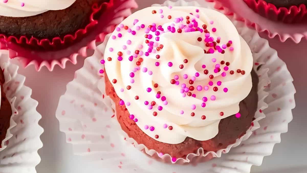 A chocolate cupcake with swirl of white frosting, topped with pink and purple sprinkles. The cupcake is in a red liner, placed on a white ruffled paper, surrounded by similar cupcakes.