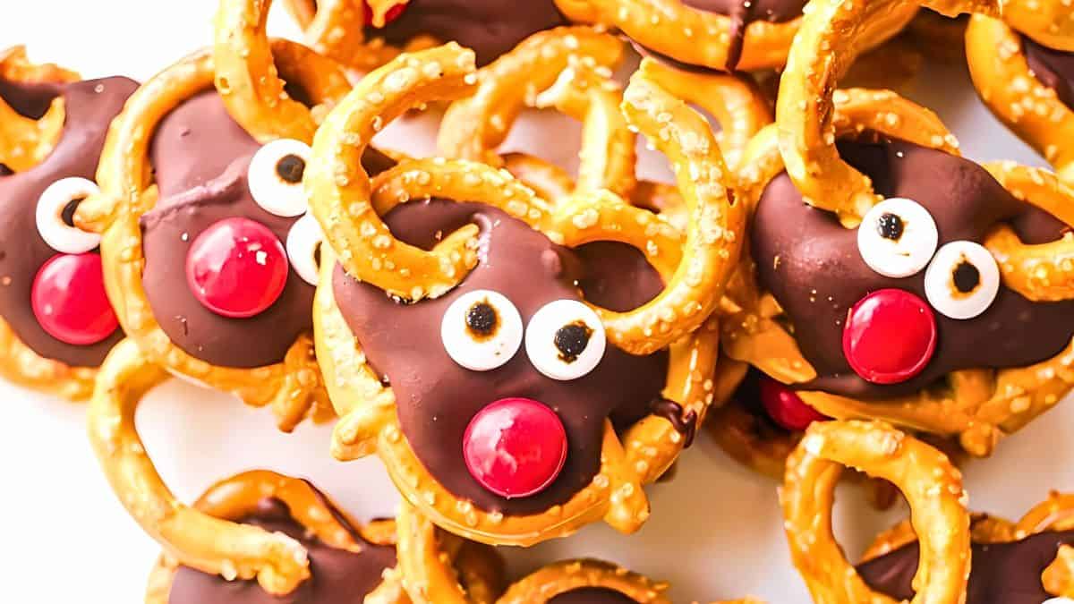 A festive snack features reindeer faces made from chocolate-covered pretzels. Each reindeer has candy eyes and a red candy nose, with pretzel antlers, arranged on a white surface.
