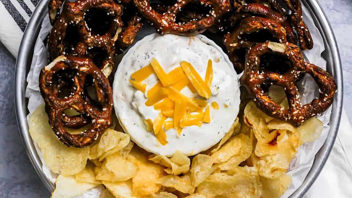 A plate with large pretzels, potato chips, and a bowl of creamy dip topped with shredded cheese.