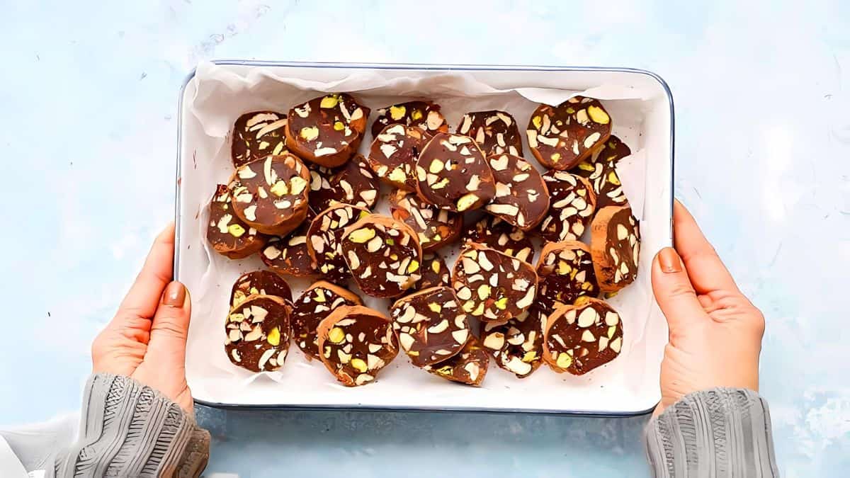 A person holding a white tray filled with chocolate-covered cookies sprinkled with chopped nuts. The tray is lined with parchment paper, and the background is a light blue surface.