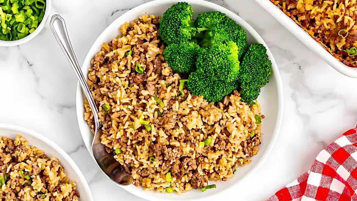 A white plate filled with seasoned ground beef, rice, and chopped green onions, topped with fresh broccoli. A fork is placed on the side, and a red and white checkered cloth is partially visible in the corner.