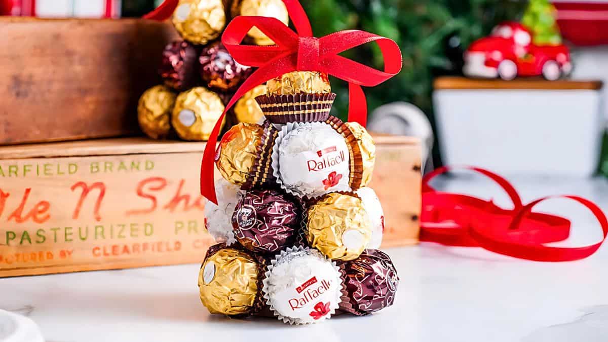 A pyramid stack of wrapped candies sits on a table, adorned with a red ribbon bow. The chocolates are in gold, white, and brown wrappings. In the background, there are wooden boxes and a small toy car.