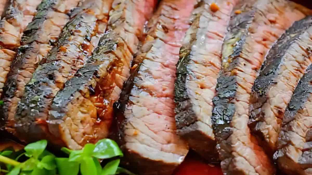 Slices of grilled steak arranged on a surface, showing a juicy pink interior with dark seared edges. Fresh green herbs are visible in the lower corner, adding a touch of color to the presentation.