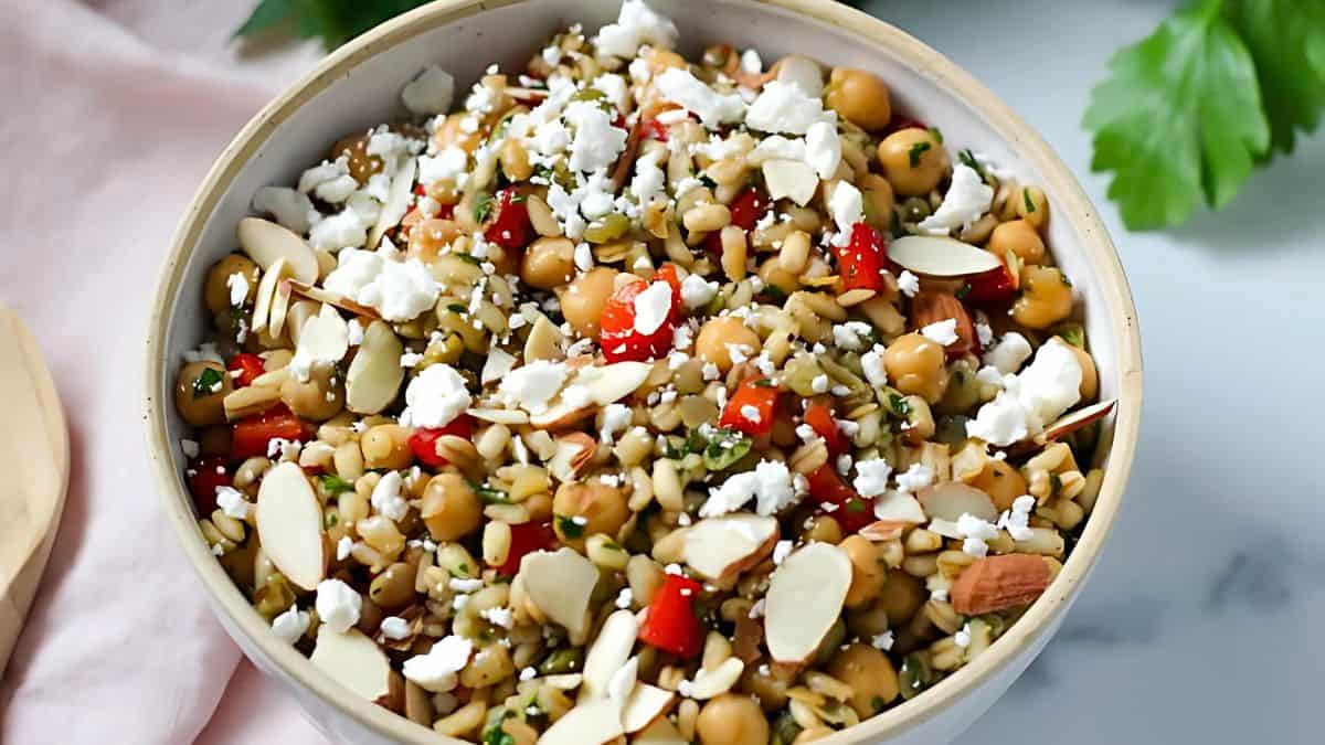 A bowl filled with a colorful salad featuring chickpeas, chopped red peppers, sliced almonds, crumbled feta cheese, herbs, and seeds. The dish is placed on a marble surface with a sprig of parsley in the background.