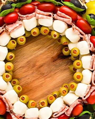 A vibrant antipasto wreath is adorned with cherry tomatoes, mozzarella balls, salami slices, black and green olives, and fresh basil leaves, arranged in a circular pattern on a wooden board.