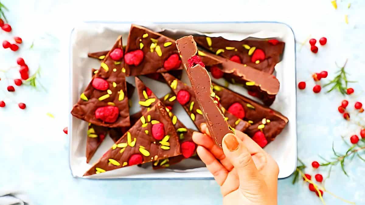 A hand holds a triangular piece of chocolate bark topped with red berries and green pistachio slivers. Other pieces are arranged in a tray. Sprigs of greenery and scattered berries are visible around the tray on a light surface.