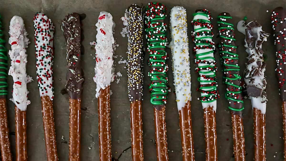 A row of pretzel rods coated in white and dark chocolate, decorated with colorful sprinkles, candy, and icing. Each pretzel features a unique design, creating a festive and decorative appearance on a dark surface.