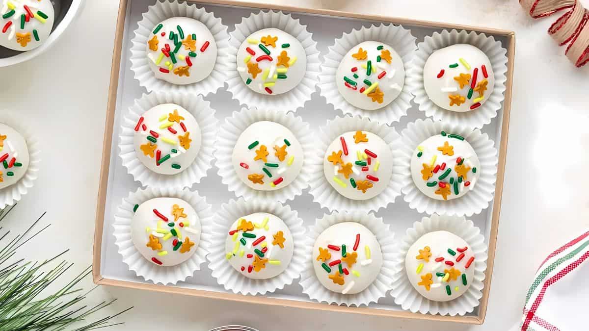 A box of 12 white chocolate truffles decorated with colorful sprinkles and star-shaped confetti, each placed in a white paper cup. The box is on a white surface, surrounded by holiday-themed decor.