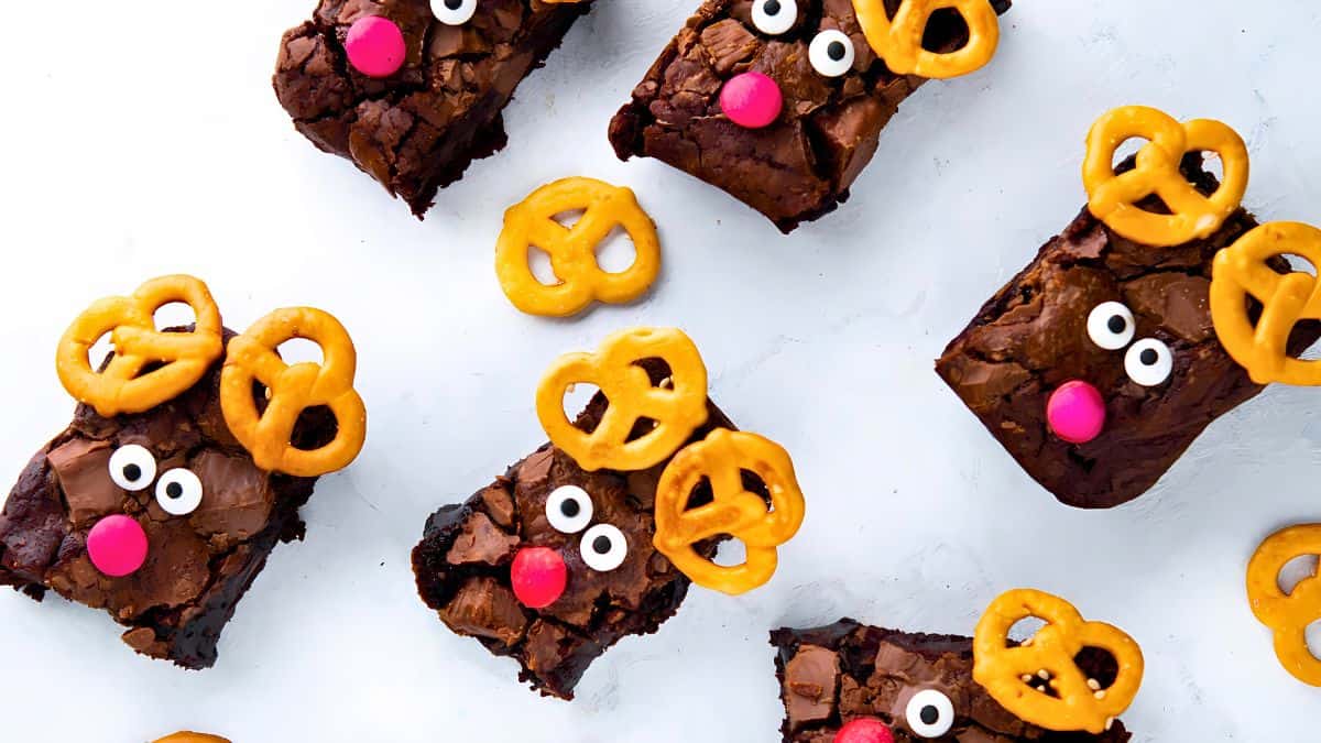 Brownies decorated as reindeer with pretzel antlers, candy eyes, and red candy noses are arranged on a white surface.