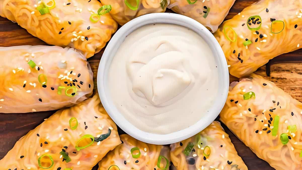 A plate of golden-brown spring rolls arranged in a circle around a bowl of creamy dipping sauce. The rolls are garnished with sesame seeds and chopped scallions, placed on a wooden surface.