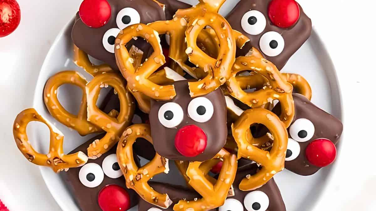 A plate of festive reindeer snacks made from chocolate-covered treats. Each has pretzel antlers, candy eyes, and a red candy nose, resembling Rudolph. The arrangement is playful and holiday-themed.