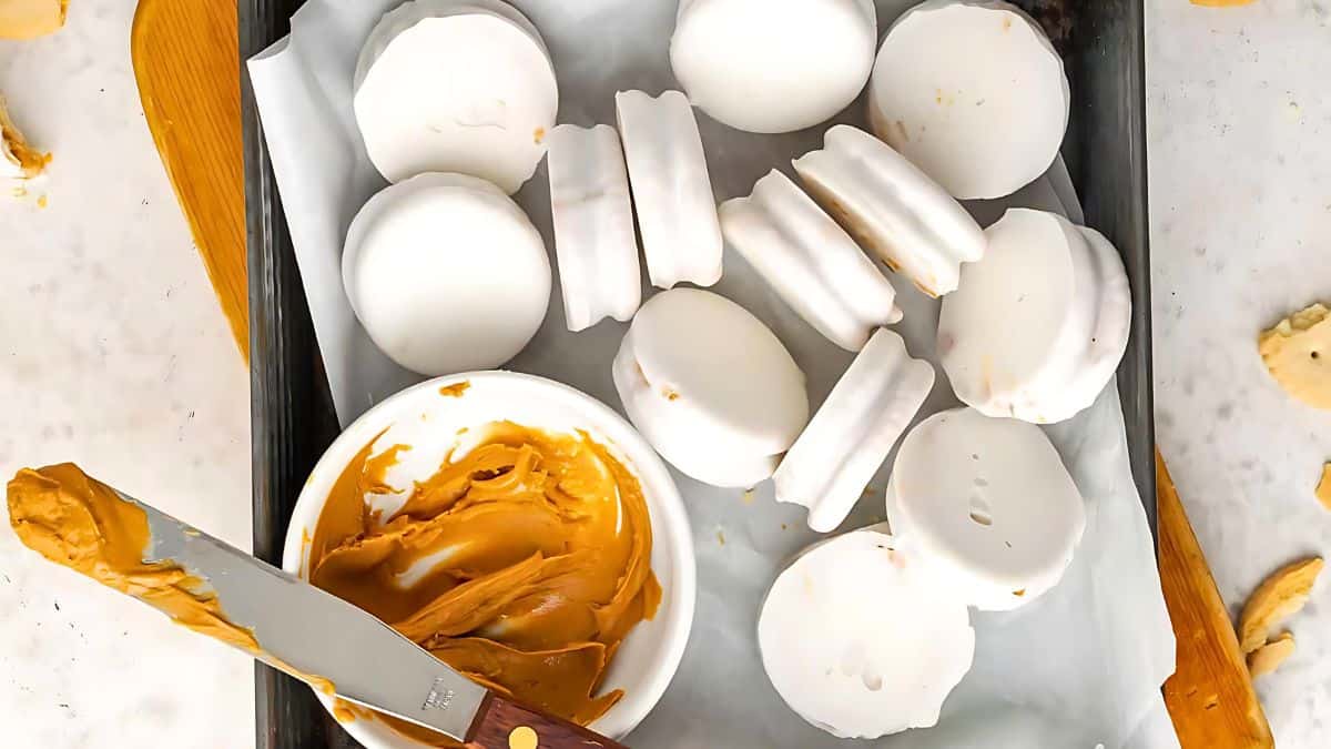 A baking tray lined with parchment paper holds white chocolate-coated cookies. A bowl of creamy peanut butter with a spreading knife rests on the tray, suggesting the cookies are being filled or topped with the peanut butter.