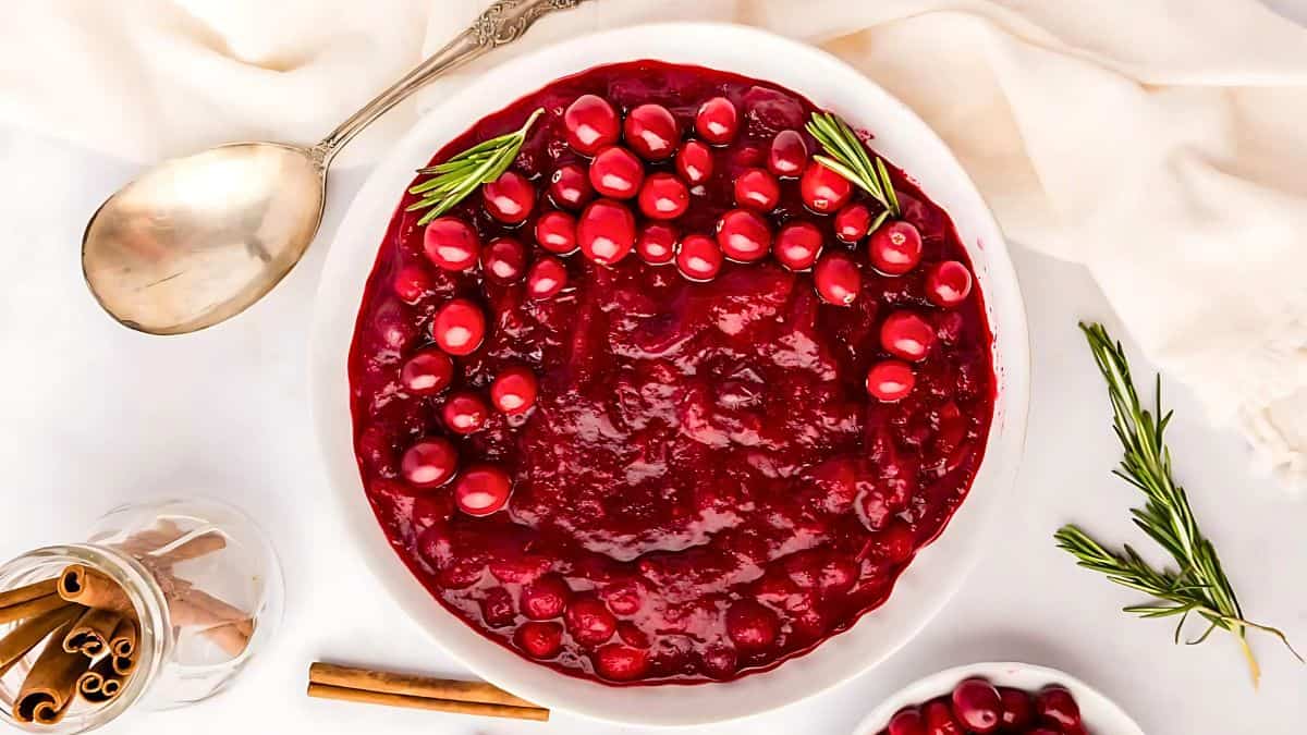 A bowl of vibrant cranberry sauce garnished with fresh cranberries and rosemary sprigs. A silver spoon and a jar of cinnamon sticks lie nearby on a white tablecloth.