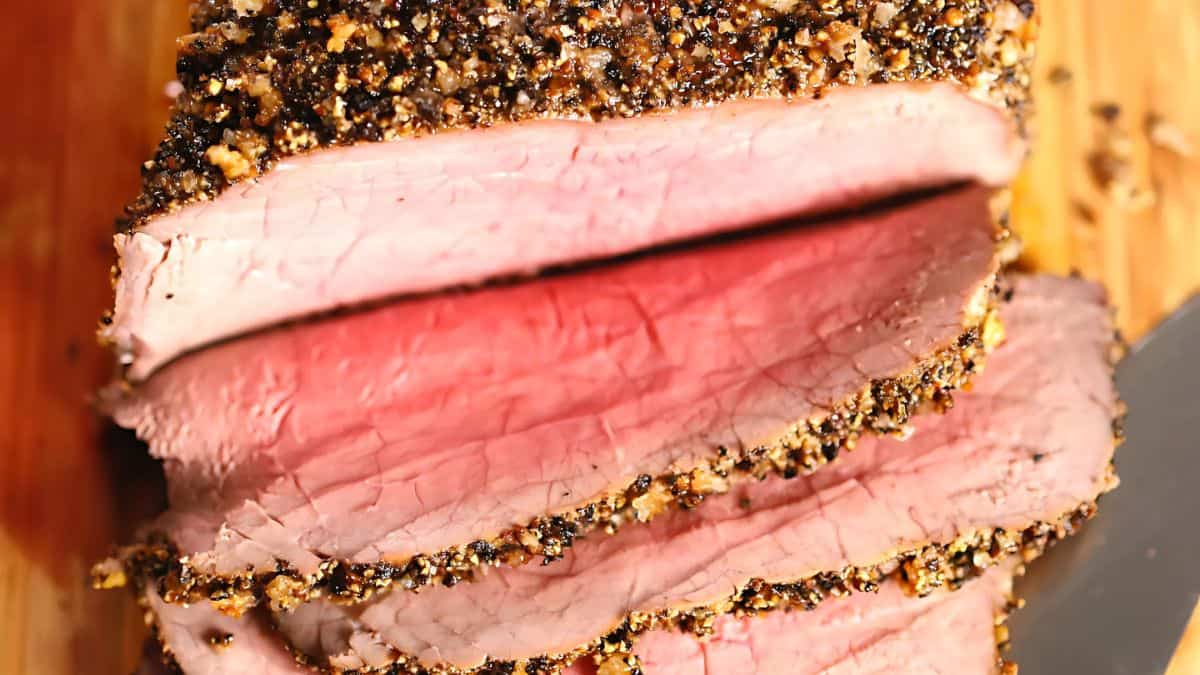Close-up of sliced roast beef coated with a pepper and spice crust on a wooden cutting board. The meat appears tender and pink in the center with a crispy, seasoned exterior. A knife is partially visible on the side.