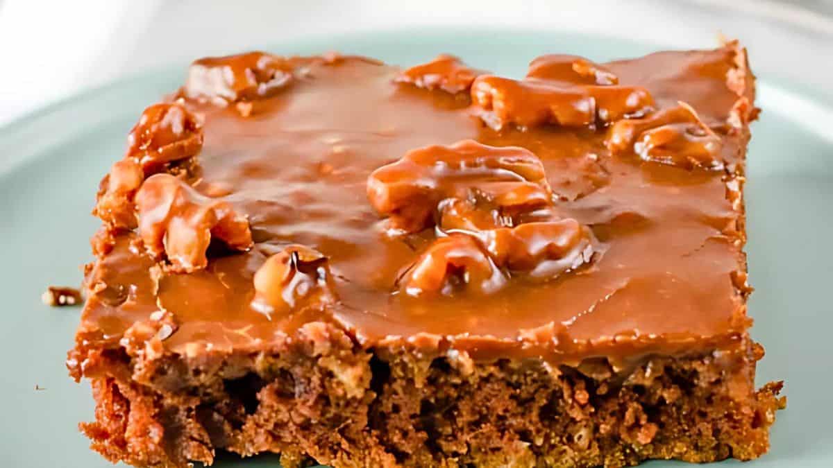 A close-up of a slice of Texas sheet cake, topped with a shiny chocolate glaze and walnuts. The cake is moist and rich in texture, displayed on a light green plate.