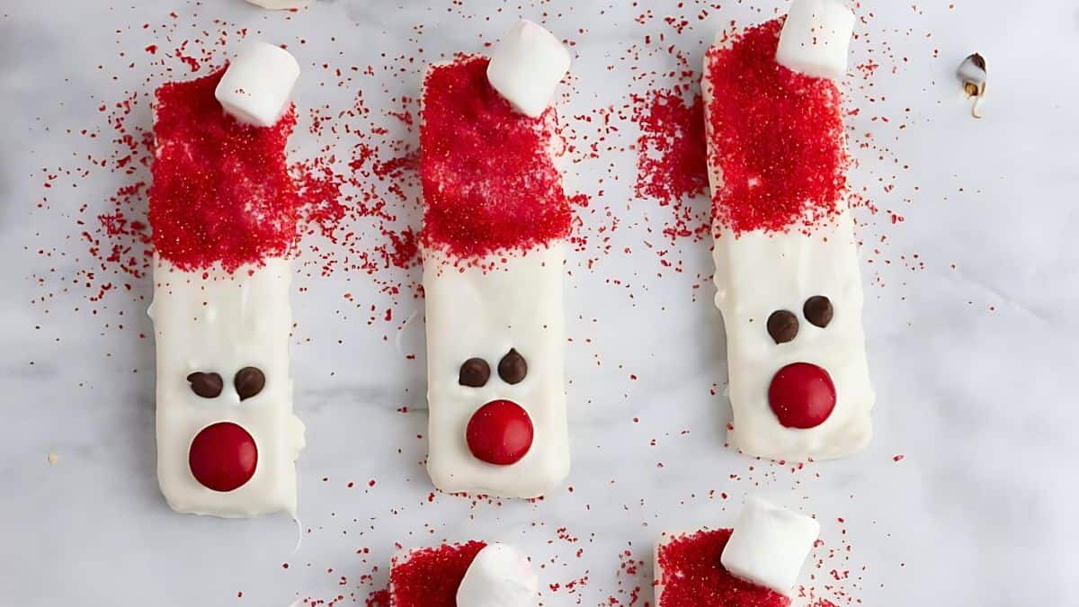 Three Santa-themed treats made with chocolate-covered cookies are decorated with red sprinkles, marshmallows for hats, brown candy eyes, and red candy noses.