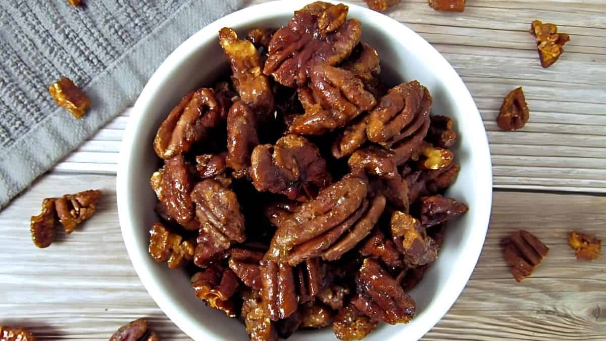 A white bowl filled with glazed pecans sits on a wooden table. A few pecans are scattered around the bowl, and a textured gray cloth is partially visible in the top left corner.