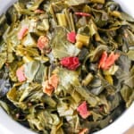 A bowl of cooked collard greens with chunks of bacon, served on a table with a white napkin and wooden utensil. The greens are vibrant and mixed with small pieces of red bacon.