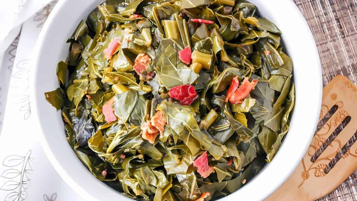 A bowl of cooked collard greens with chunks of bacon, served on a table with a white napkin and wooden utensil. The greens are vibrant and mixed with small pieces of red bacon.
