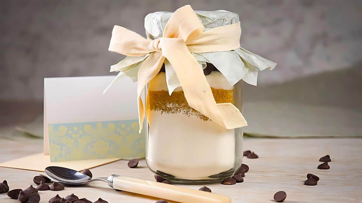 A jar filled with layered dry baking ingredients is decorated with a cream-colored ribbon and a paper cover. It sits next to a blank card and spoon, surrounded by scattered chocolate chips on a light surface.