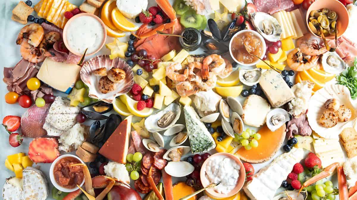 A vibrant charcuterie board filled with assorted cheeses, meats, seafood, fruits, nuts, and dips. The display includes shrimp, clams, grapes, olives, strawberries, and an array of sliced cheeses and cured meats, artfully arranged.