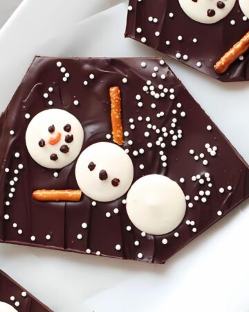 Chocolate bark decorated with snowman designs made from white candy discs and pretzel sticks on a white platter. The snowmen have chocolate chip eyes and buttons, with white sprinkles scattered around. A decorative snowflake is partially visible.