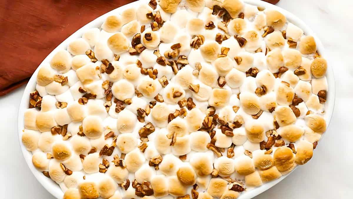 Oval dish filled with baked sweet potatoes, topped with golden-brown marshmallows and sprinkled with chopped pecans.