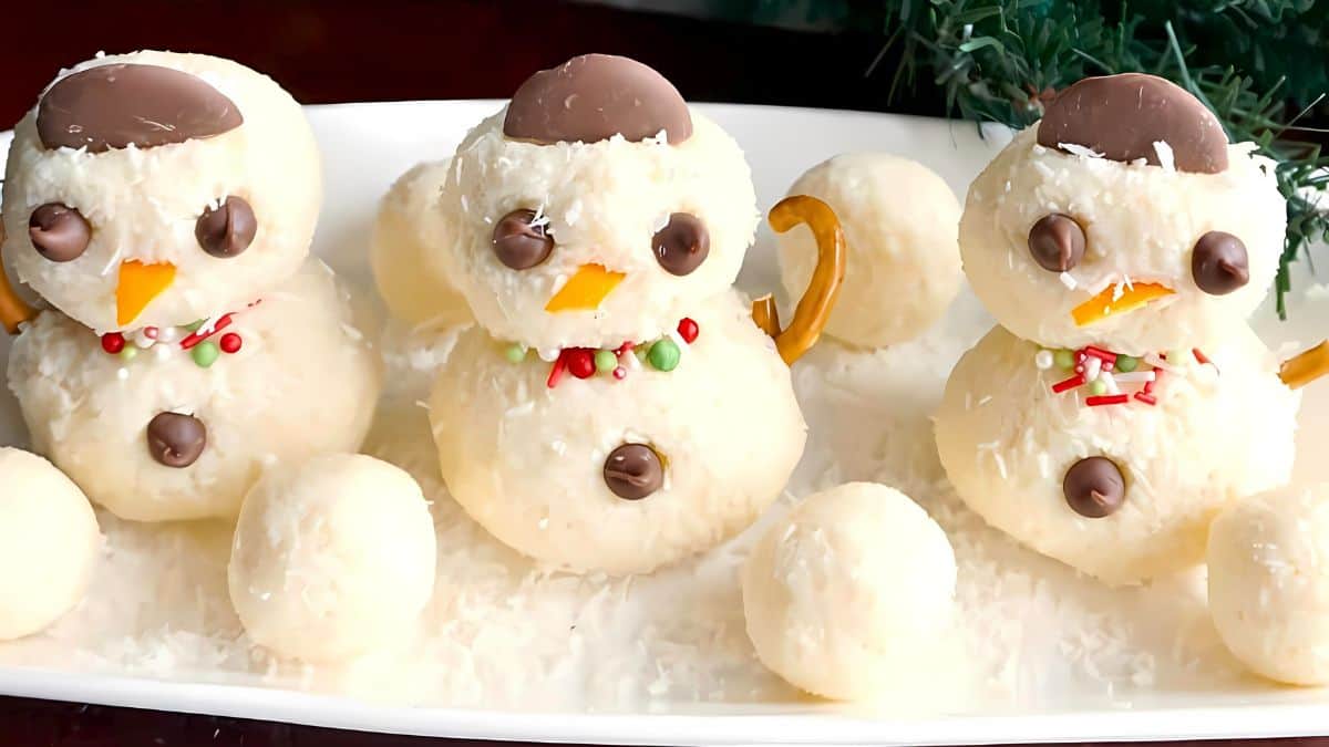 Three snowman-shaped coconut desserts on a white platter. Each snowman has chocolate eyes, an orange candy nose, and a pretzel piece as a scarf. Greenery borders the background.