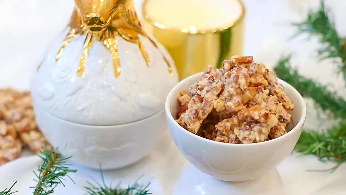 A bowl of nut clusters sits on a white table beside a white and gold decorative container. Greenery surrounds the setup, and a blurred candle is in the background, creating a festive and cozy atmosphere.